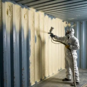 Contractor applying closed-cell spray foam insulation to the steel interior walls of a shipping container home, showing code-compliant R-value application.